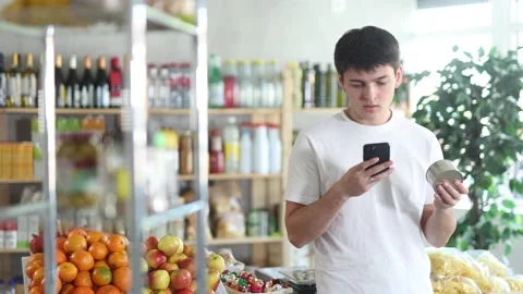 Young man customer at shop scans QR code on tin cans of preserves using phone Stock Footage 309184030