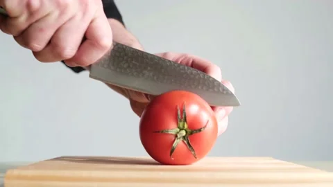 A young man cuts a ripe red tomato in half on a wooden board. Stock Footage 133808885