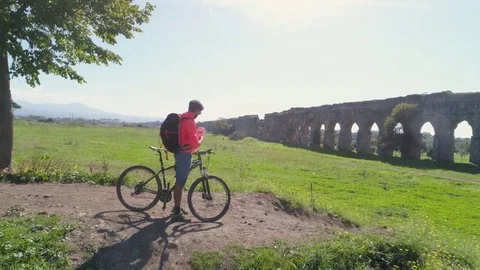 Young man cyclist checking his bike on dirt road in front of ancient roman Stock Footage 84736463