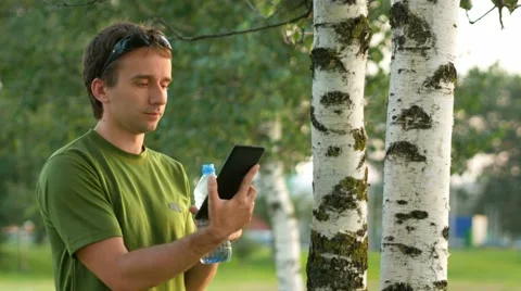 A young man cyclist touch tablet near the birch and drink water from a bottle Video stock 66454285