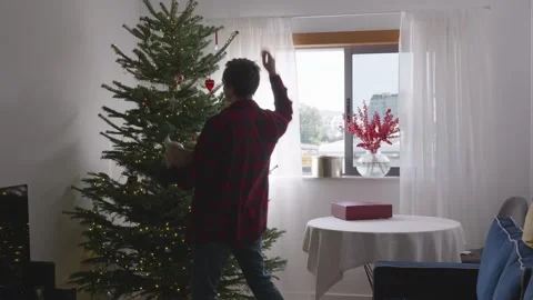 Young man dancing and decorating Christmas tree in cozy bright living room Stock Footage 324923888