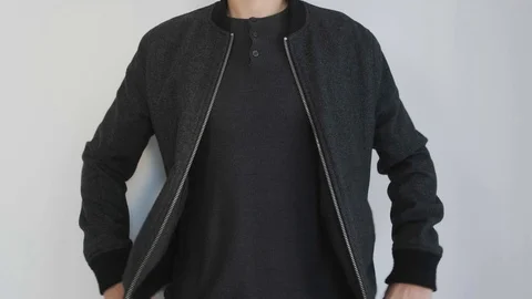 A young man in dark clothes zips up his jacket standing near a white wall. Stock Footage 121761963