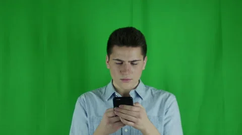Young man is distracted on phone and looks up annoyed in front of a greenscreen Vidéo 59083911