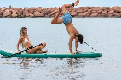 Young man doing a handstand on a paddle board while woman sitting watching hi Stock Photos