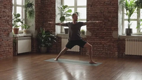 Young man doing warrior yoga pose in studio with wooden floor and big windows Video stock 107103293