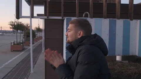 A young man in a down jacket sits on a bench near the port and warms his hands 스톡 동영상 123208763