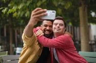 Young Man With Down Syndrome And His Mentoring Friend Sitting And Taking Selfie Stock Photos