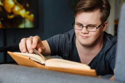 Young man with down syndrome reading book while lying on couch Stock Photos