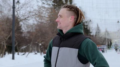 Young man with dreadlocks walking through snowy urban park, beaming while Stock Photos