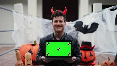 A young man dressed as a vampire devil with makeup holds a laptop with a green Stock Footage 250410173