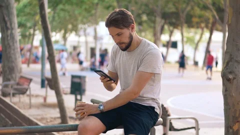 Young man drinking beer and checking smartphone in park sitting on rail Stock Footage 111778616