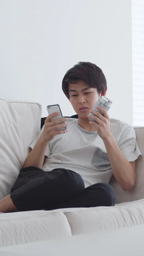 Young man drinking beer while looking at smartphone on couch Video stock 161922301