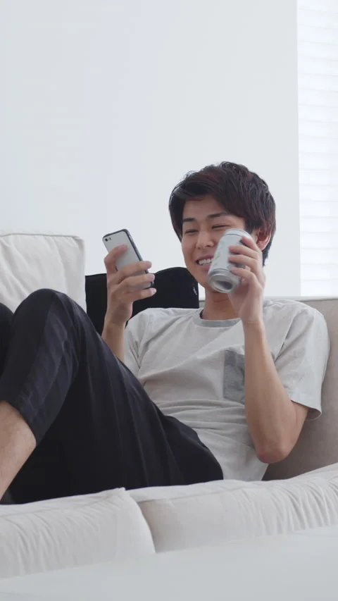Young man drinking beer while looking at smartphone on couch Stock Footage 161922331