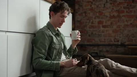 Young man is drinking coffee and touching dog while sitting on floor in home Stock Footage 198484026
