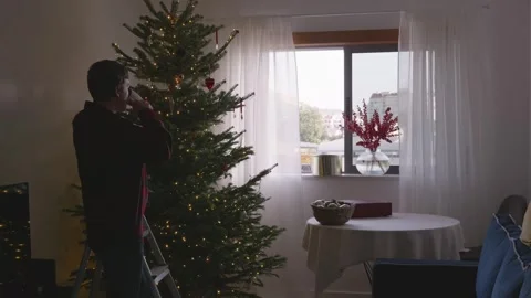 Young man drinking coffee and looking at decorated Christmas tree in cozy room Stock Footage 324923794