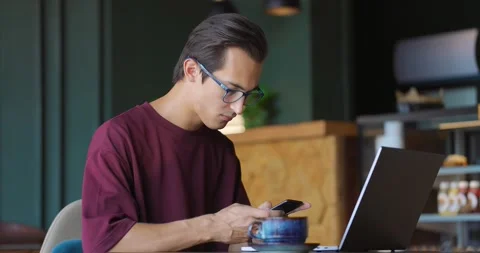Young man drinking coffee in a cozy cafe while typing on laptop and chatting Stock Footage 163996565