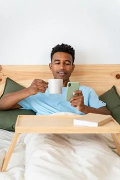 Young man drinking a cup of coffee and using a telephone in bed. Stock Photos