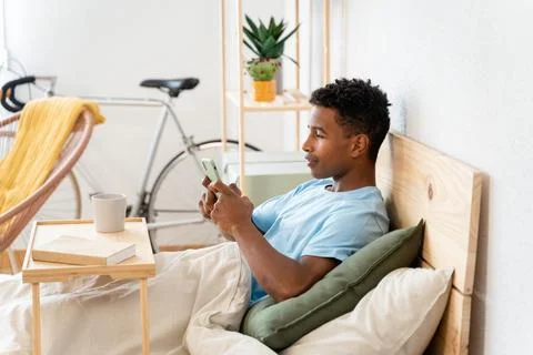 Young man drinking a cup of coffee and using a telephone in bed. Stock Photos