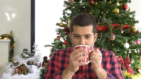 Young man drinking hot chocolate in a red mug in front of Christmas tree Stock Footage 140673029