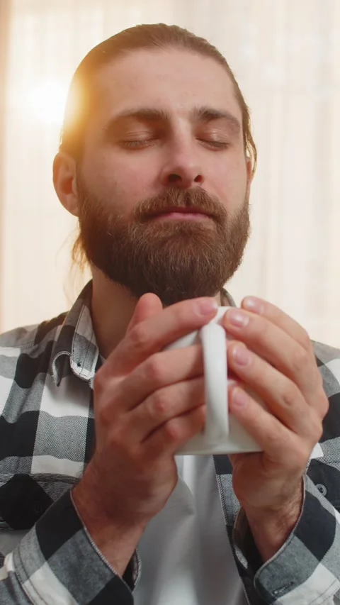 Young man drinking hot drink at home sofa, aroma, eyes closed, content sip Stock Footage 348058527