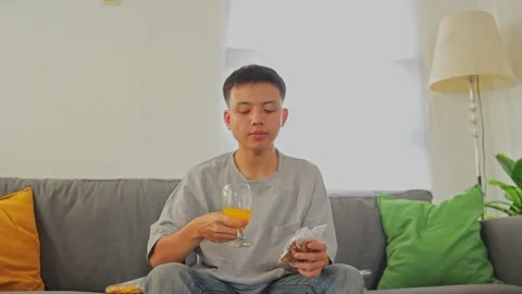 Young man drinking juice and broad beans while sit on cozy sofa Stock Footage 346264940