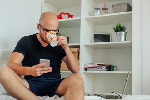 Young man is drinking something from mug and using mobilephone. Home backgrou Stock Photos