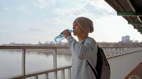 Young Man is Drinking Water from Bottle on the Bridge over the River and City Vidéo 74250184