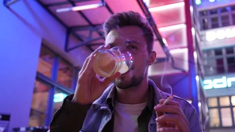 Young Man drinks Beer and smokes Cigarette in a Night Street Bar Outdoors Stock Footage 139869875