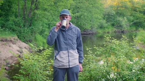 A young man drinks tea from a thermos mug near the forest river Stock Footage 90957774
