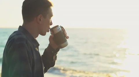 Young man drinks from a thermocup on the beach slow motion Stock Footage 54635892