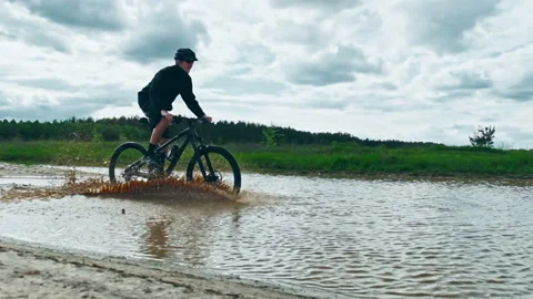 Young man drives into a large puddle on a black mountain bike. Cyclist rides Stock Footage 135915373