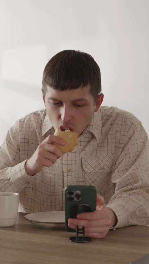 Young man eating breakfast snack and scrolling smartphone at kitchen table Stock Footage 335082340