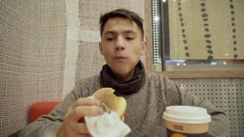 A young man is eating a hamburger in a cafe. Video stock 82517587