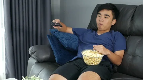 Young man eating popcorn while sitting on a couch at home and watching TV Stock Footage 93414884