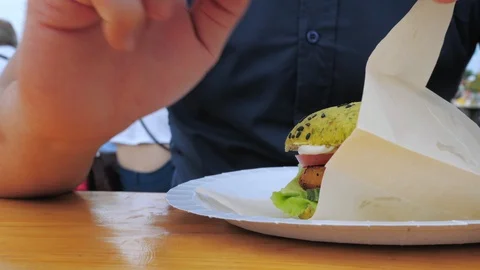 A young man eats a hamburger or cheeseburger with a green bun in an open-air Stock Footage 113351332