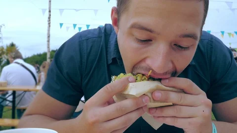 A young man eats a hamburger or cheeseburger with a green bun in an open-air Video stock 113351971
