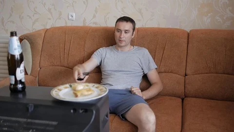 Young man eats near the TV and switches the channels on the TV. Stock Footage 69815512