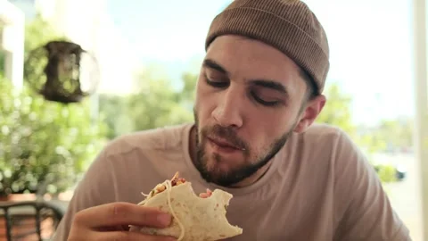 A young man eats tacos while sitting in a sunny outdoor cafe, enjoying his meal Stock Footage 342545195