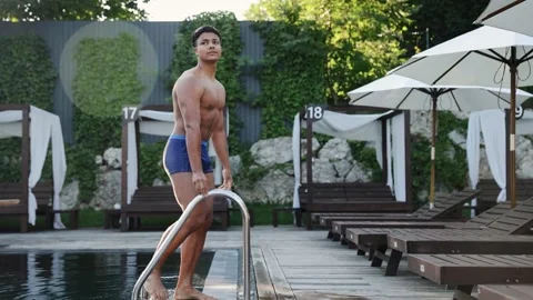 Young man emerging from swimming pool stepping onto wooden deck at luxury resort Stock Footage 328160148