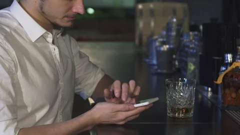 Young man enjoying a drink while texting on his smartphone at a stylish bar Stock Footage 311941631