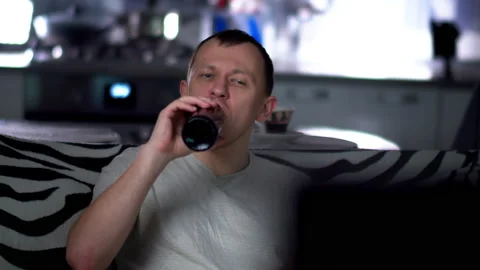 A young man in the evening sitting on the sofa watching TV and drinking beer Stock Footage 132829324