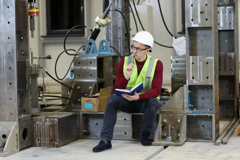 Young man, factory engineer in red pullover and white hard hat holding notebo Stock Photos