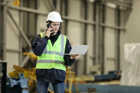 Young man, factory engineer in a work dress and white hard hat holding smartp Stock Photos