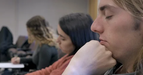 Young man falling asleep in a classroom while his classmates work on assignments Stock Footage 105910046