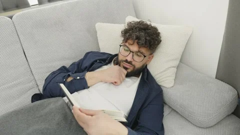 Young man falling asleep while reading book on the sofa Stock Footage 345128539