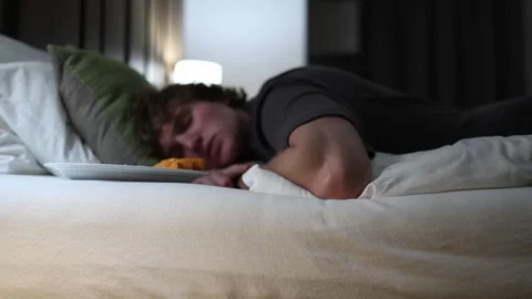 Young man falls asleep on bed beside plate with food in dim hotel room light sho Stock Footage 318217760