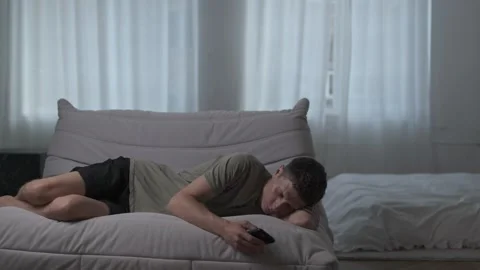 Young man falls asleep scrolling on his phone on a sofa in a minimalist Stock Footage 311443000