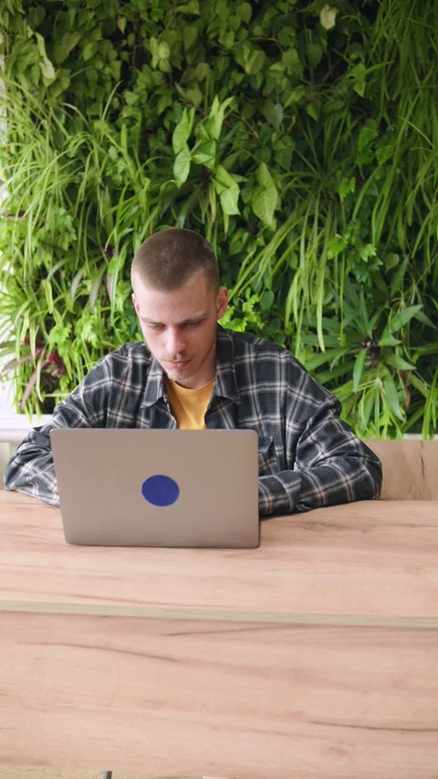 A young man is focused on his laptop while sitting at a wooden table. He is in a Stock Footage 346151434