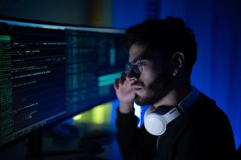 Young man focused on programming in a dim workspace, glowy screens display .. 库存照片