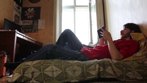 A young man with a gadget lying on sofa in his room (dolly shot) Video stock 82211254